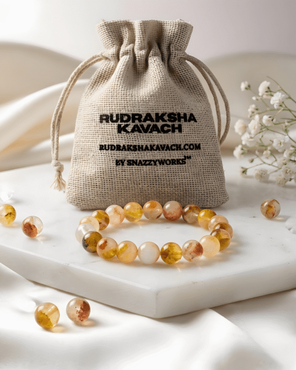 A circular beaded bracelet made of polished citrine gemstones in warm shades of pale yellow, gold, and reddish-brown, resting on a white hexagonal stone slab. The elegant setting features softly draped cream-colored fabric in the background, a sprig of delicate white baby's breath flowers, and a few small, clear crystals scattered around the base.